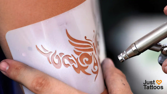 Semi-permanent tattoo being applied with airbrush tool over stencil on skin showing design before 24-hour development